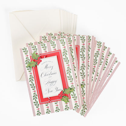 A stack of Mistletoe Striped Boxed Cards (10 ct), featuring “Merry Christmas and Happy New Year” on the front with holly accents and red borders, sits beside matching white envelopes. These festive cards add a charming touch to your holiday greetings.
