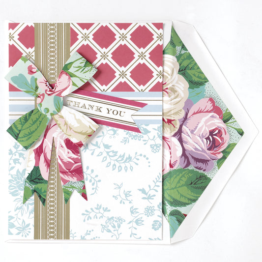 The Amelie Thank You Card features a pink and white floral design with roses and a decorative bow, paired with a matching printed lined envelope with a rose motif. The partially open envelope reveals the 5" x 7" card.