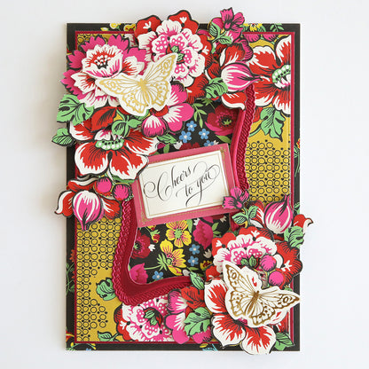 The Hermosa Multi Cultural Cardstock is a colorful greeting card showcasing vibrant floral patterns inspired by Hispanic culture, intricate cutout butterflies, and a central message that reads "Cheers to you." This card is embellished with various textured designs and crafted from high-quality cardstock.
