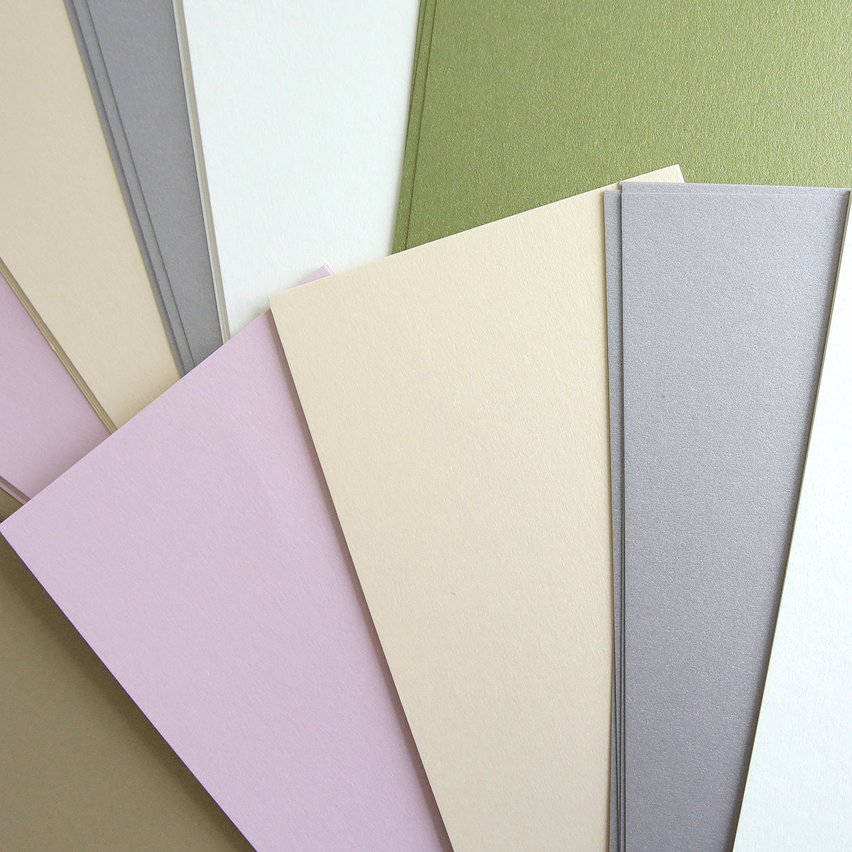 The Luxury Shimmer Cardstock - Antique, featuring a range of pastel hues such as beige, pink, green, and gray, is elegantly displayed fanned out on a flat surface.