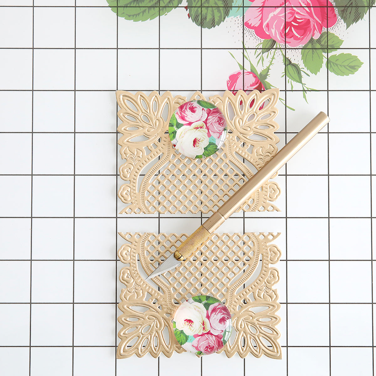 A gold craft knife lies on two ornate beige floral paper cutouts atop the Glass Magnetic Craft Mat 16 x 22, which features a checkered surface and a rose print in one corner.