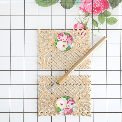 A gold craft knife lies on two ornate beige floral paper cutouts atop the Glass Magnetic Craft Mat 16 x 22, which features a checkered surface and a rose print in one corner.