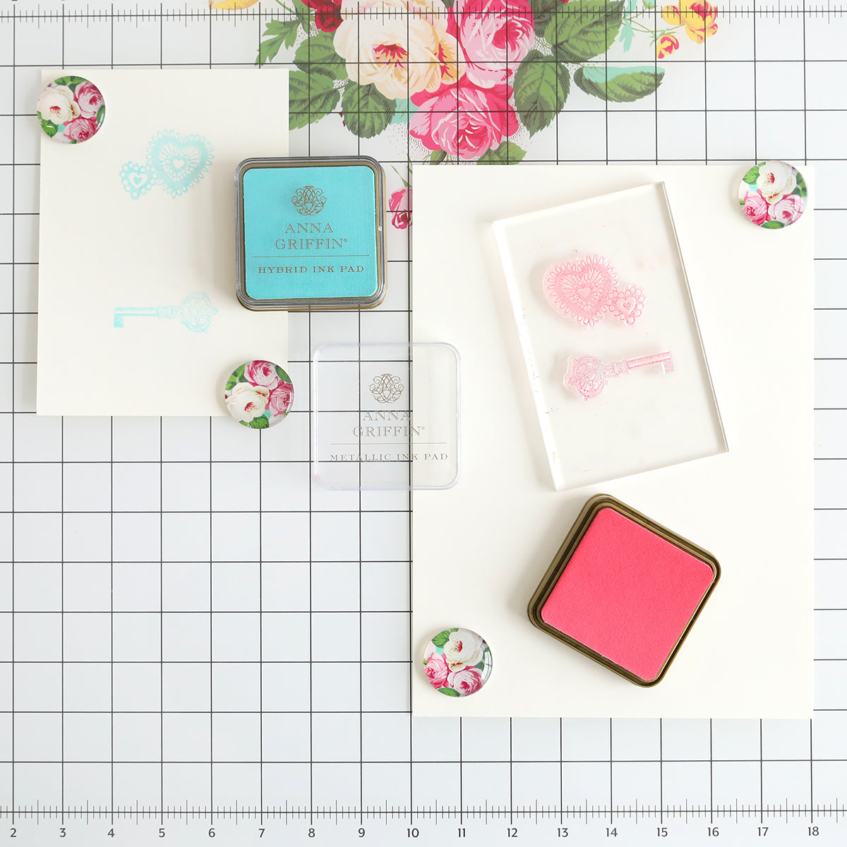 Two ink pads, a clear stamp block, decorative magnets, and stamped designs are displayed on the Glass Magnetic Craft Mat 16 x 22, which features a white grid background and a floral design motif at the top center.