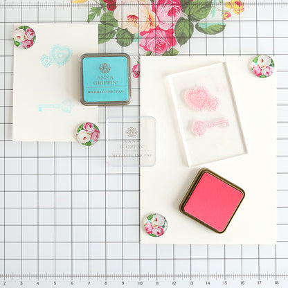 Two ink pads, a clear stamp block, decorative magnets, and stamped designs are displayed on the Glass Magnetic Craft Mat 16 x 22, which features a white grid background and a floral design motif at the top center.