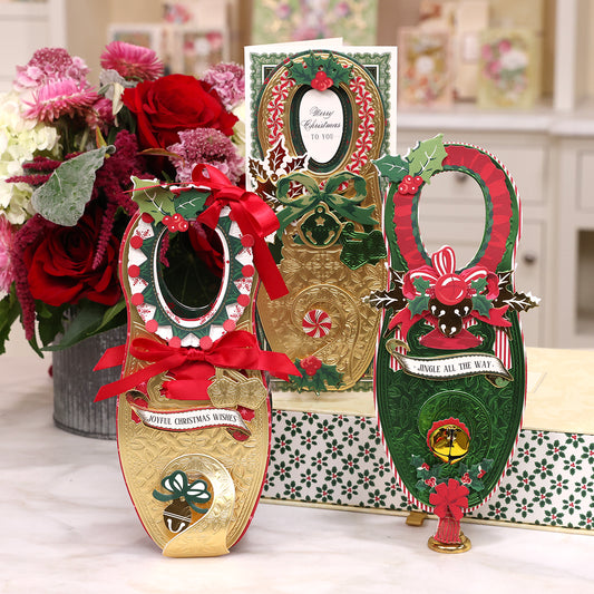 Three elaborately decorated items from the Paper Elf Shoes Finishing School Class, featuring festive elements like ribbons, holly leaves, and candy canes, arranged with a floral bouquet in the background on a white surface.