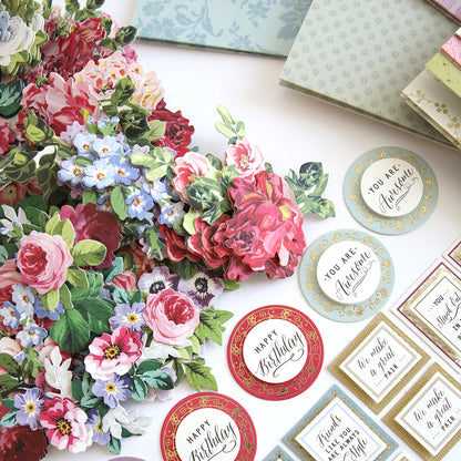 An assortment of floral decorations, layers of various greeting cards, and a Paper Sneakers Refill Kit spread out on a white surface.