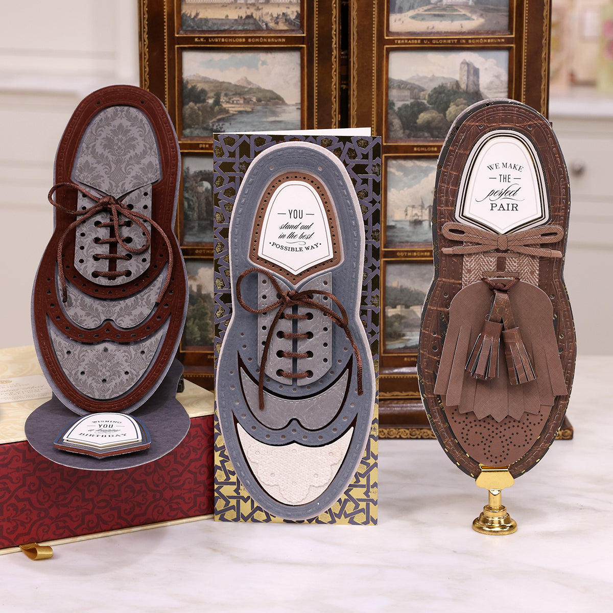 Three shoe-shaped greeting cards are displayed upright on a table. Each card is intricately designed to resemble a different style of dress shoe, with detailed patterns and messages inside, looking as if they belong in a Paper Wingtips Finishing School Class.