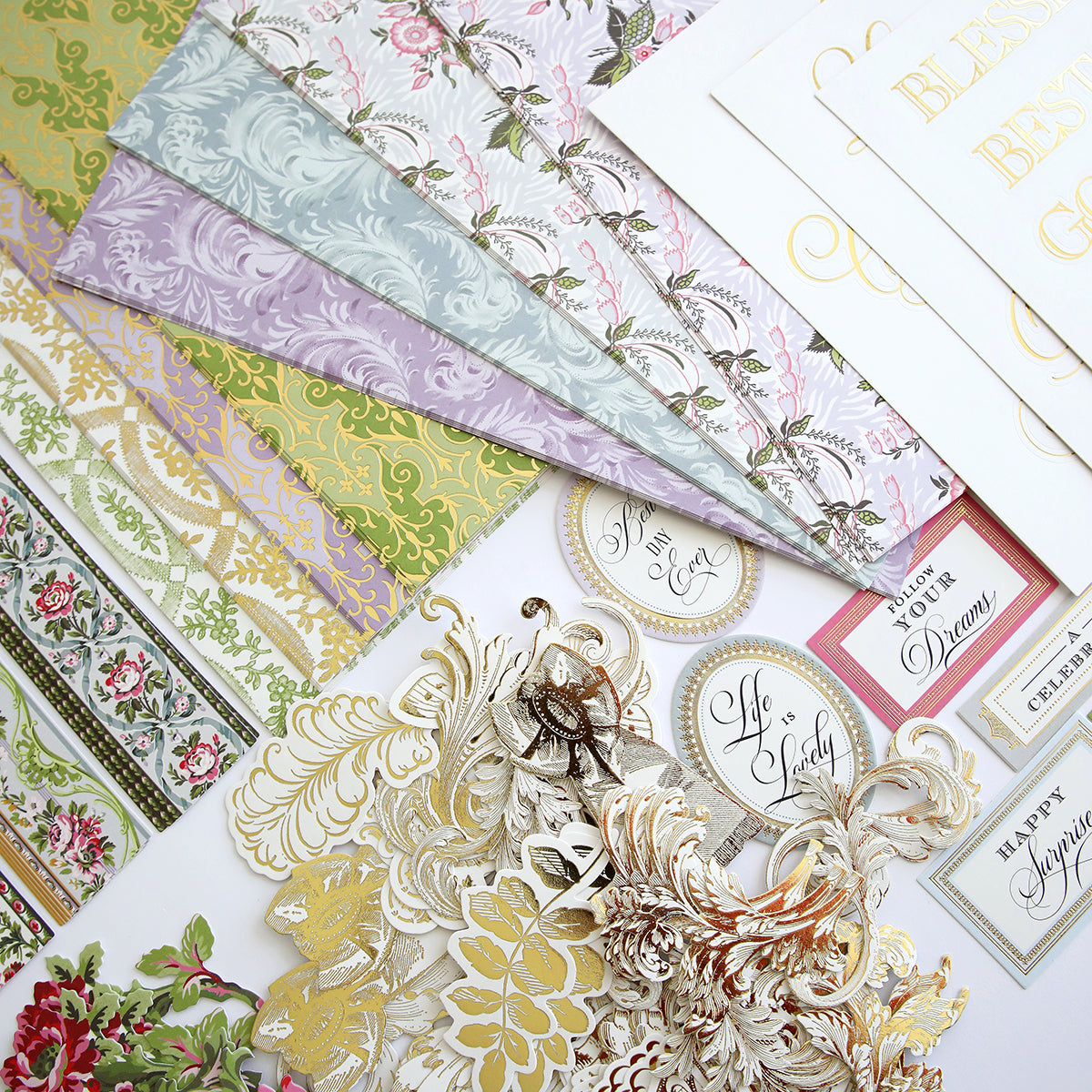 An assortment of Phoebe Papers and Embellishments, ornate die-cuts, and gold-accented greeting cards arranged on a surface.