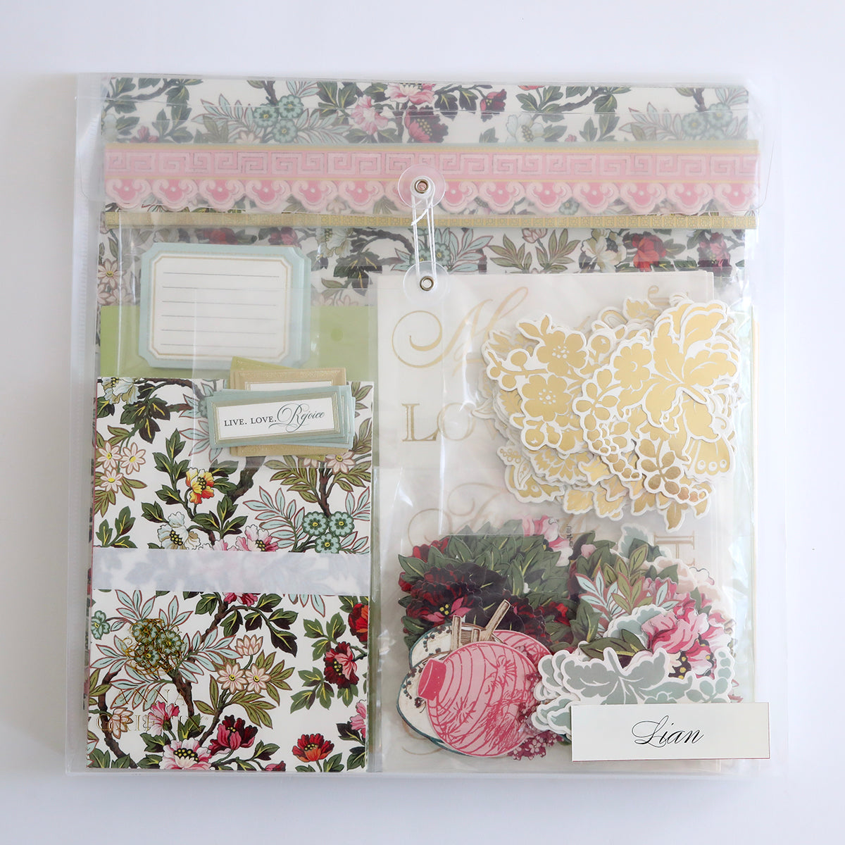 The Crop at Home 5 Ultimate Materials Kit comes in a clear plastic pouch with floral-patterned scrapbook stationery items, die-cut flowers, tags, labels, and cardstock cards neatly arranged inside.