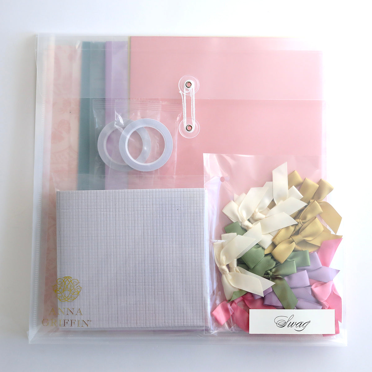 The Create Crop At Home 5 Papercraft and Scrapbook Event kit is perfect for scrapbooking, with pastel sheets, tape rolls, and a labeled "Swag" clear bag of multicolored ribbons. Ideal for Anna Griffin events or any papercrafting occasion.