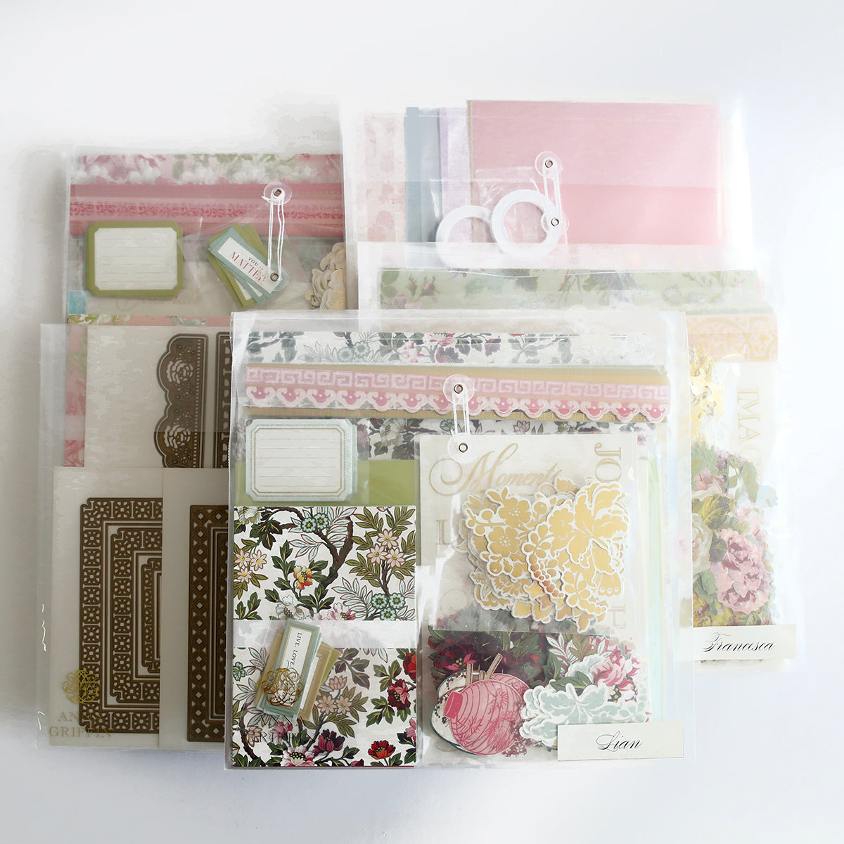 The Crop at Home 5 Ultimate Materials Kit features clear plastic packets with floral and decorative scrapbook supplies such as paper, tags, frames, die-cut shapes, and embellishments, organized on a white surface.