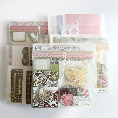 The Crop at Home 5 Ultimate Materials Kit features clear plastic packets with floral and decorative scrapbook supplies such as paper, tags, frames, die-cut shapes, and embellishments, organized on a white surface.