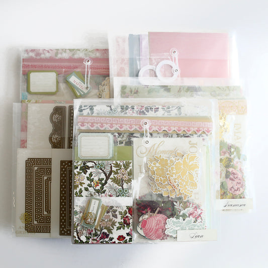 The Crop at Home 5 Ultimate Materials Kit features clear plastic packets with floral and decorative scrapbook supplies such as paper, tags, frames, die-cut shapes, and embellishments, organized on a white surface.