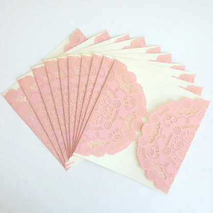 The Pink Lace Diecut Gatefold 10 pack features several cream-colored envelopes, each adorned with a pink paper lace doily, fanned out on a white surface, reminiscent of elegant wedding or diecut invitations.