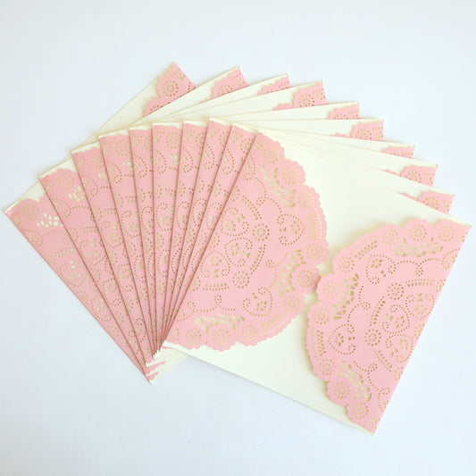 The Pink Lace Diecut Gatefold 10 pack features several cream-colored envelopes, each adorned with a pink paper lace doily, fanned out on a white surface, reminiscent of elegant wedding or diecut invitations.
