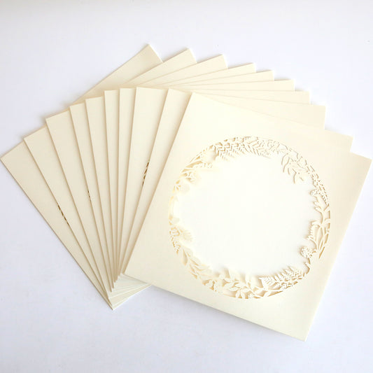 A fan-shaped arrangement of Ivory Lasercut Circle Sleeve 10 pack, featuring square, cream-colored invitation sleeves with an intricate circular floral cutout in the center, displayed on a white background.