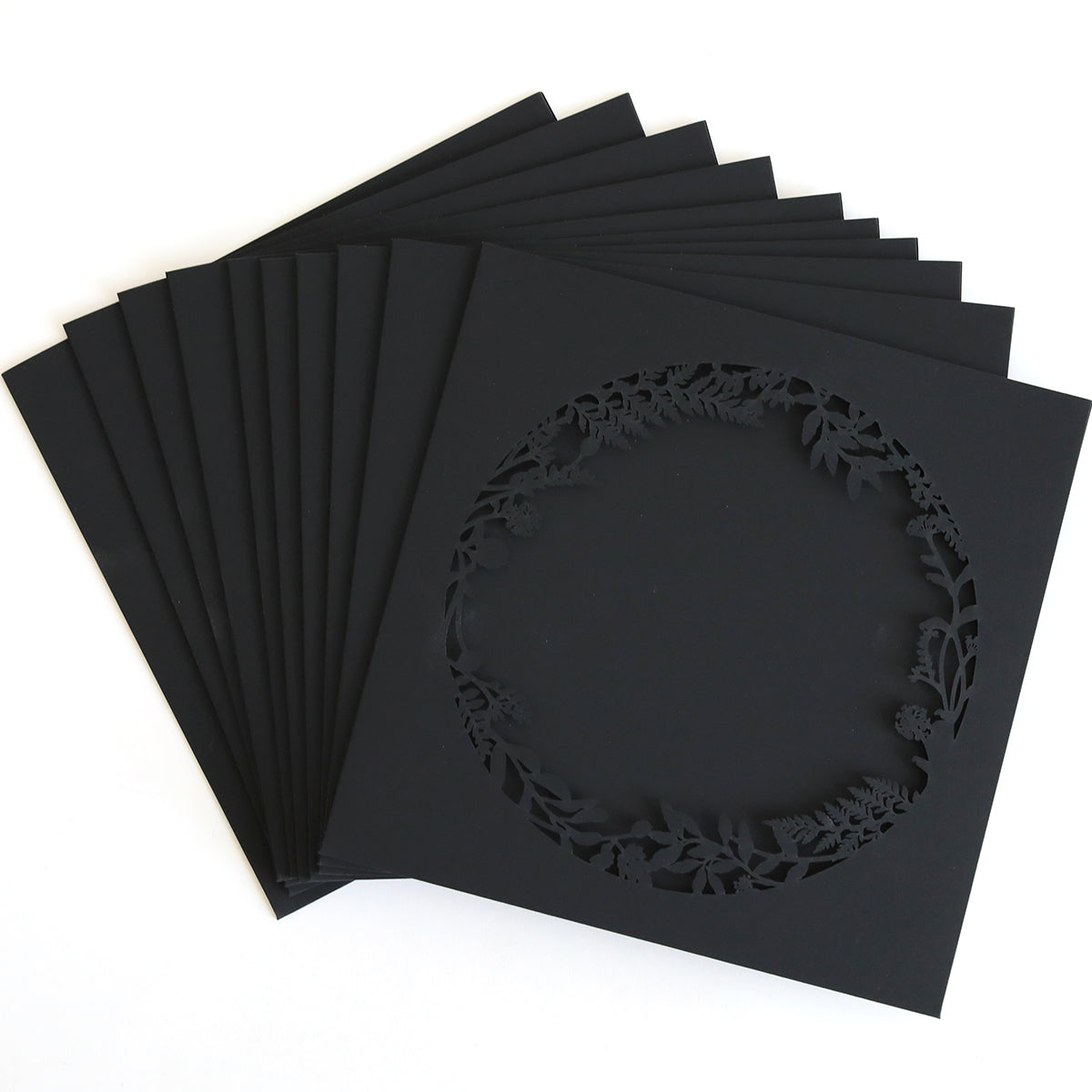 The Black Lasercut Circle Sleeve 10 pack features black square paper cards, each with a circular, laser-cut floral and leaf border—ideal for creating elegant invitation sleeves.