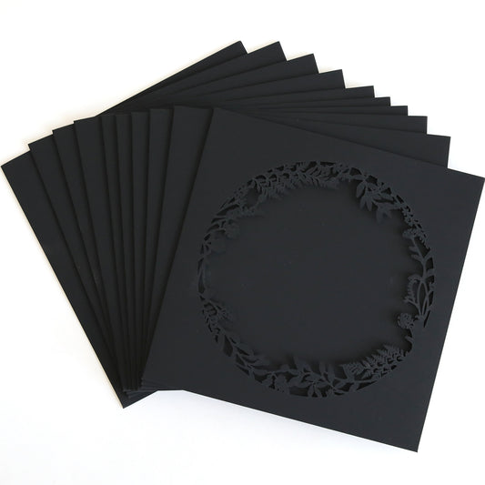 The Black Lasercut Circle Sleeve 10 pack features black square paper cards, each with a circular, laser-cut floral and leaf border—ideal for creating elegant invitation sleeves.