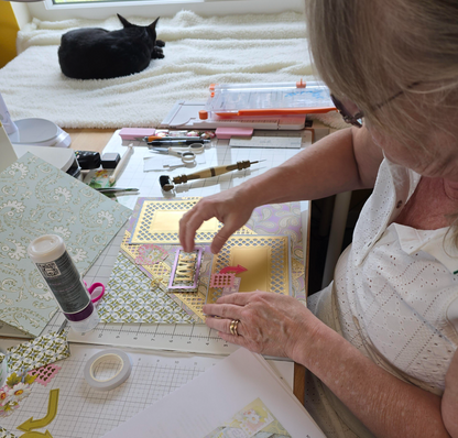 A woman crafts a scrapbook page at her table, surrounded by tools and materials from Anna Griffin | Create Fall 2026, while her black cat lounges on the windowsill—a cozy scene of creative anticipation.