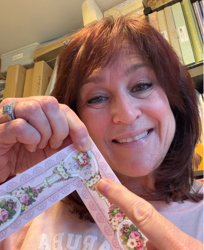 A woman smiles while holding a floral-patterned paper strip, presenting her project in a cozy room—an ideal moment from Anna Griffin | Create Fall 2026, perfect for craft classes and celebrating creativity.