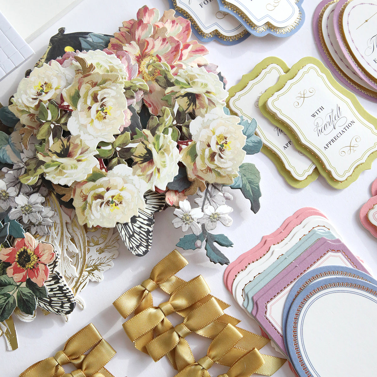 A collection of floral paper cutouts, decorative labels, and gold bows from the Simply Appreciation Easel Card Making Kit are beautifully arranged on a white surface, perfect for crafting supplies and handmade cards.