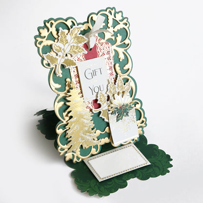 An intricately designed holiday card featuring gold holly and evergreen motifs with the text "Gift for You" on a prominent tag, standing upright with green and gold details on a white background. This exquisite creation is part of the Simply Joyful Easel Card Making Kit, perfect for fans of handmade cards.