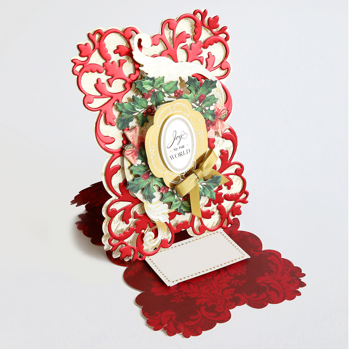 An ornate, festive pop-up greeting card featuring red and gold accents, holly leaves, and a message reading "Joy to the World." The card is open and standing upright, showcasing the elegance achievable with the Simply Joyful Easel Card Making Kit.