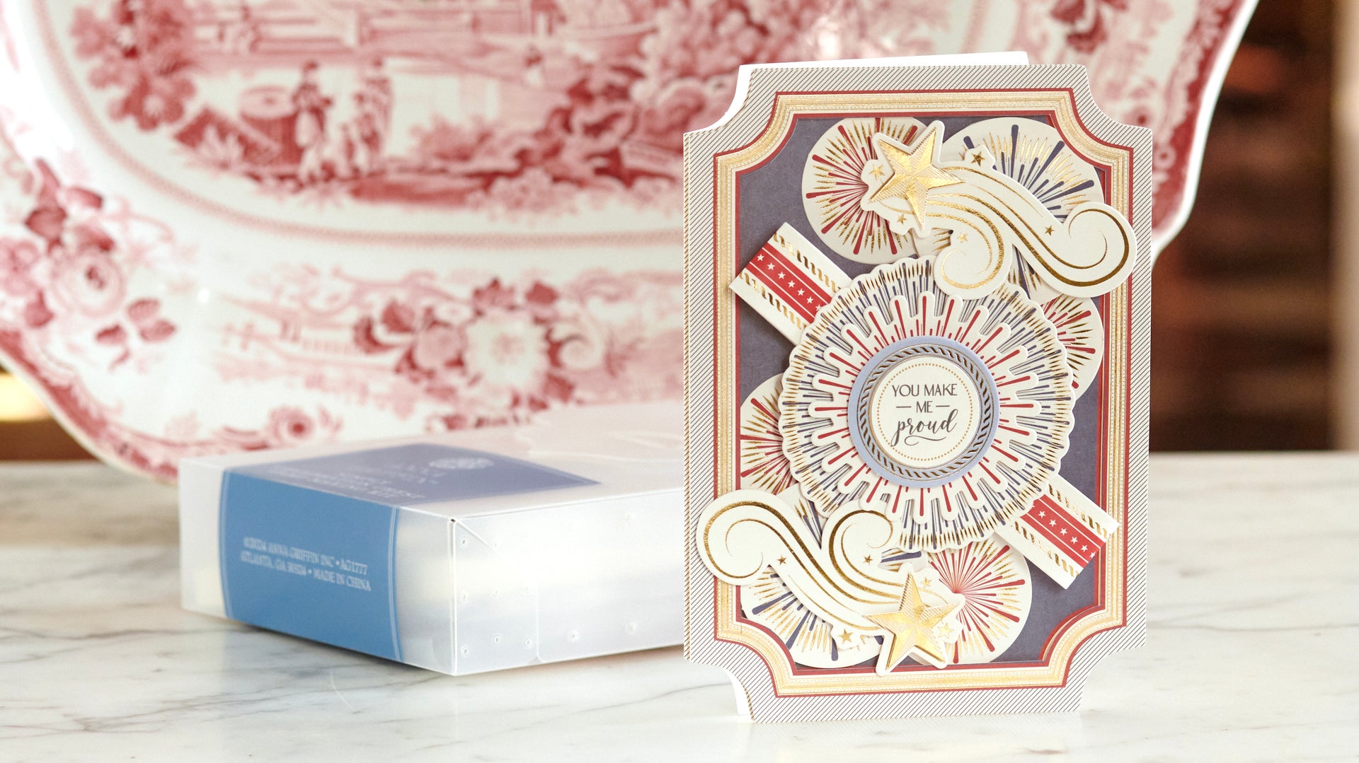 A decorative greeting card with fireworks, stars, and the message "You make me proud" stands upright on a marble surface with a red-patterned plate in the background.