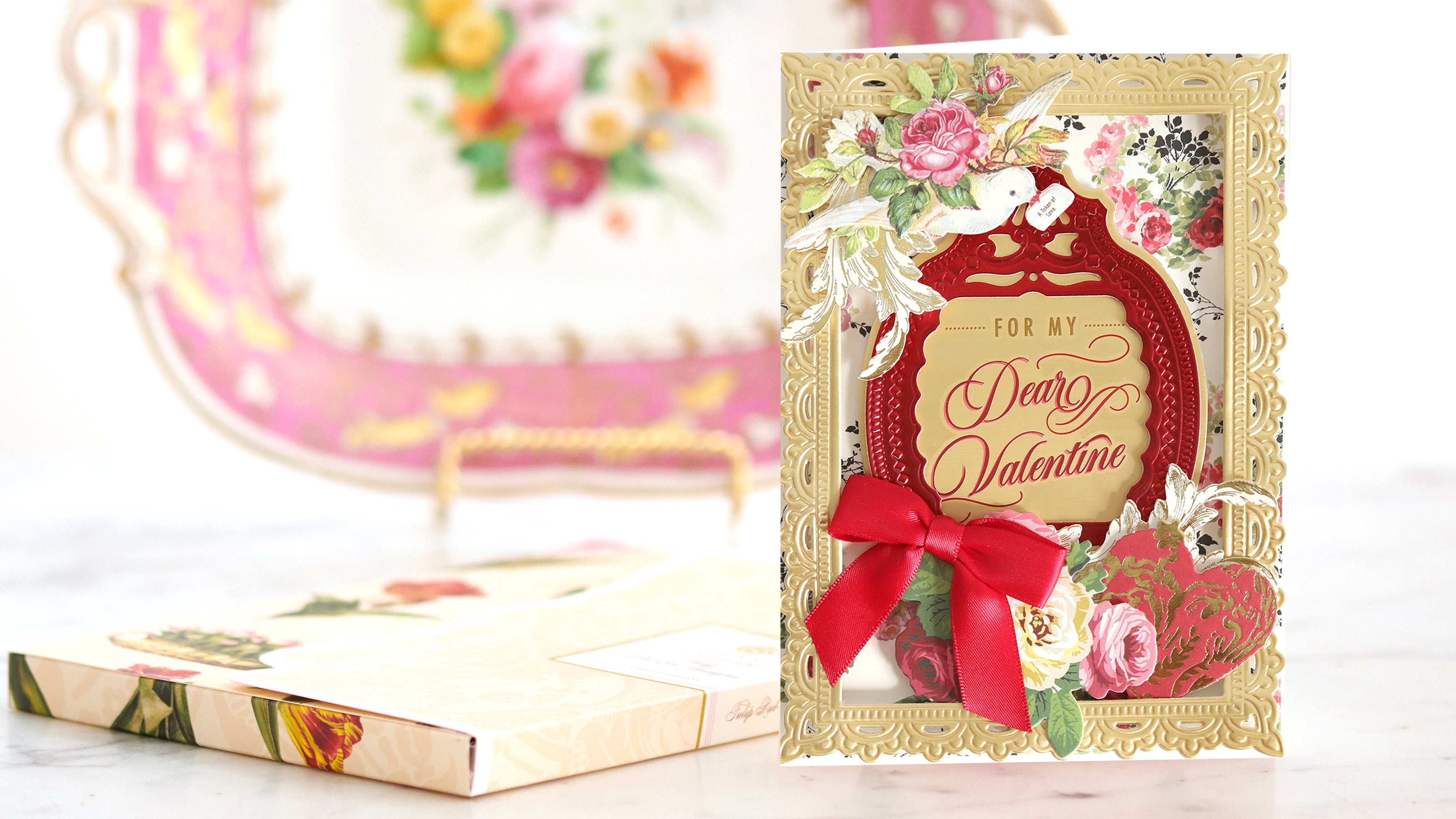 An ornate Valentine's Day card with gold and red detailing, featuring roses and a red bow, stands in front of a floral decorative plate.