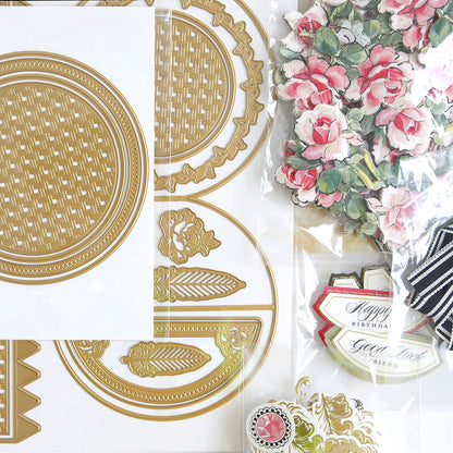 Assorted decorative craft supplies, including gold die-cut shapes and floral paper embellishments, arranged on a white surface.
