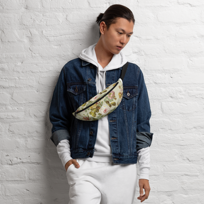 A person in a denim jacket and white pants leans against a white brick wall, sporting an eye-catching floral-patterned fanny pack with adjustable straps.