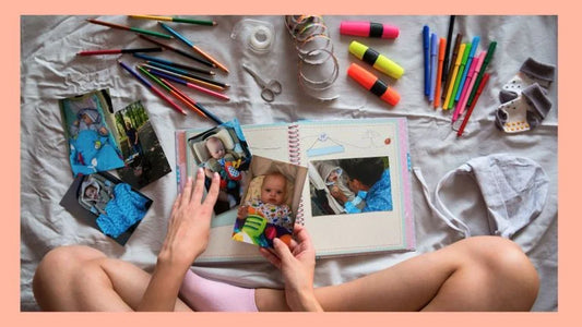A person sits cross-legged on a bed, creating a scrapbook with baby photos, colored pencils, markers, and other craft supplies scattered around.