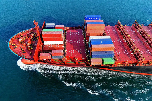Aerial view of a large red cargo ship carrying multicolored shipping containers moving through blue ocean water.
