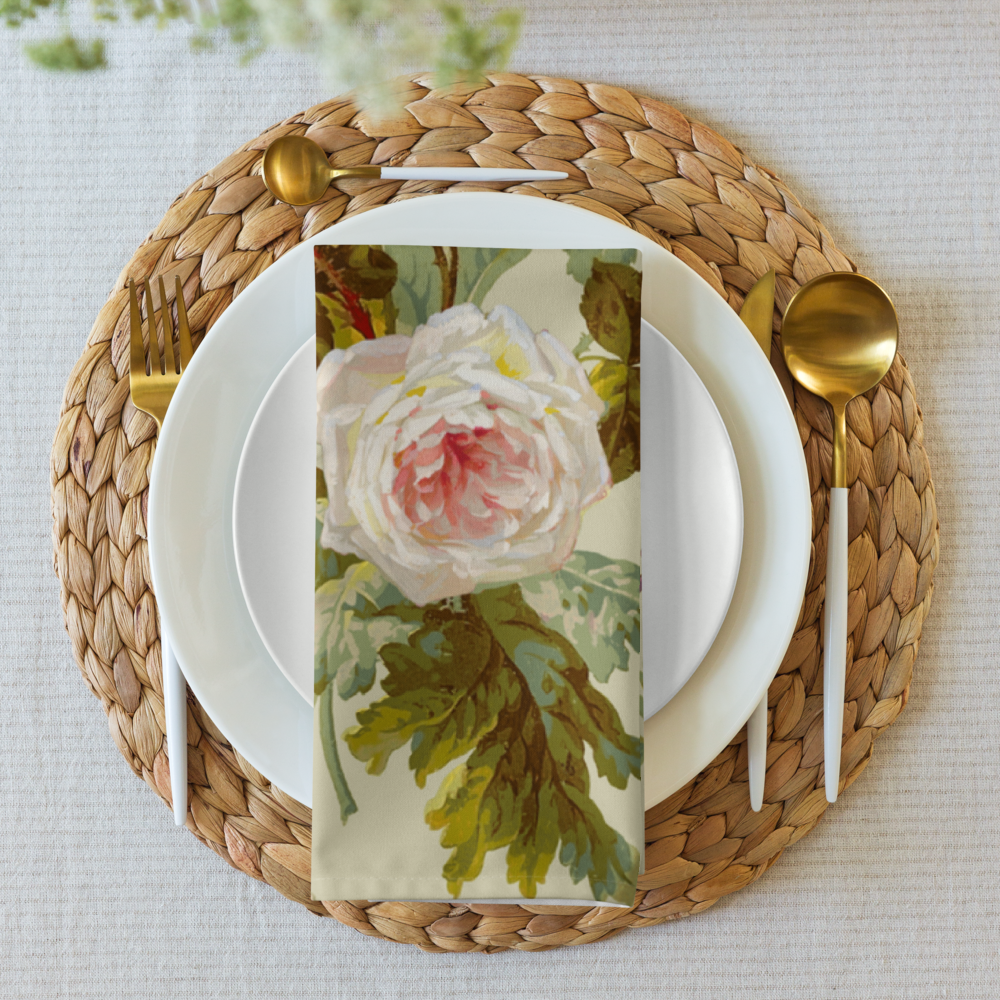 A custom napkin from the Cloth Napkin Set of 4, featuring a white and pink rose, is placed on a white plate. The setting includes a woven placemat and gold cutlery, enhancing the dining experience on a light-colored tablecloth.