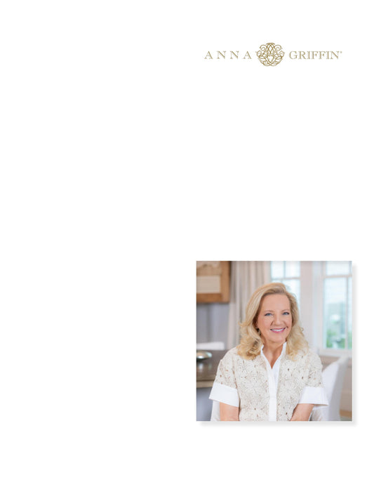 A woman with blonde hair and a white blouse is smiling while seated indoors. The logo "Anna Griffin" appears at the top right corner.