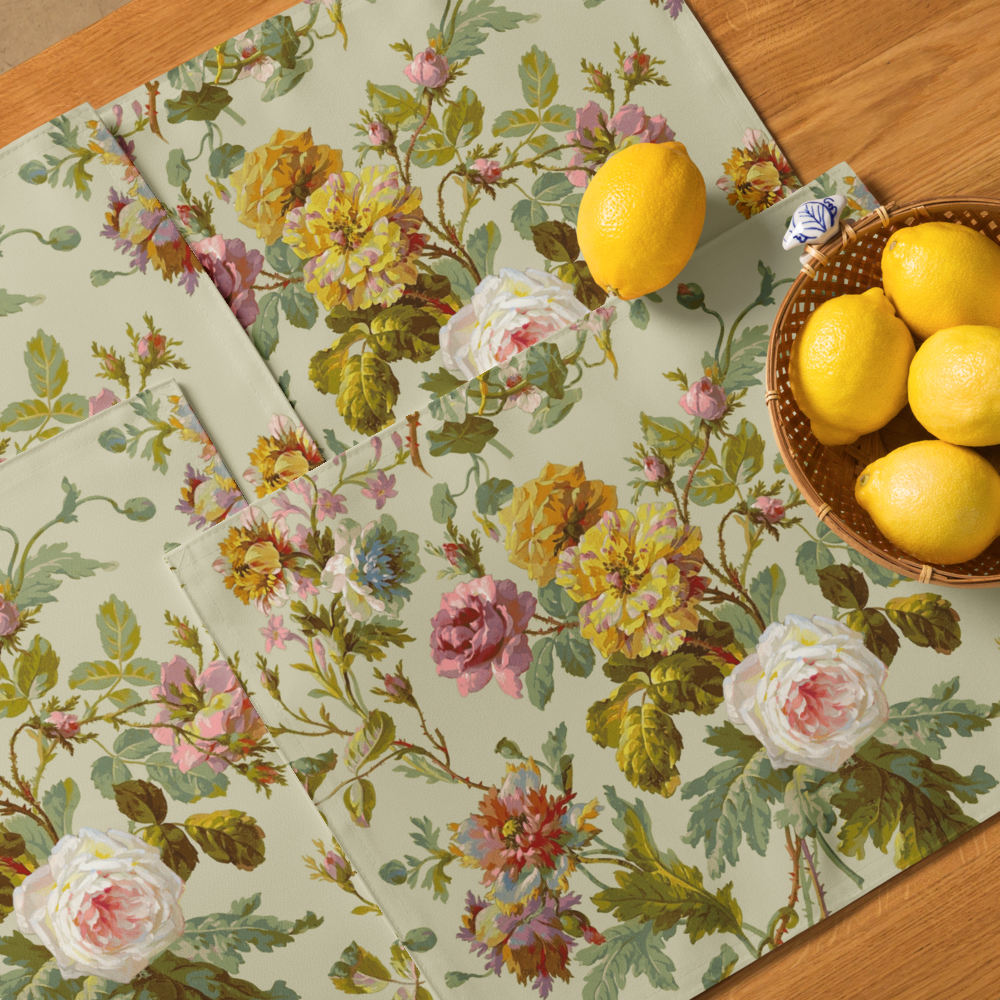 Floral napkins with rose and leaf patterns are displayed on a wooden surface alongside a Placemat Set of 4, completed by a wicker bowl filled with lemons.