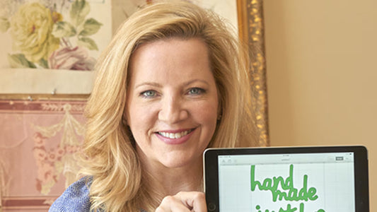 A woman smiles while holding a tablet displaying green text that reads "hand made." Floral art is visible in the background.