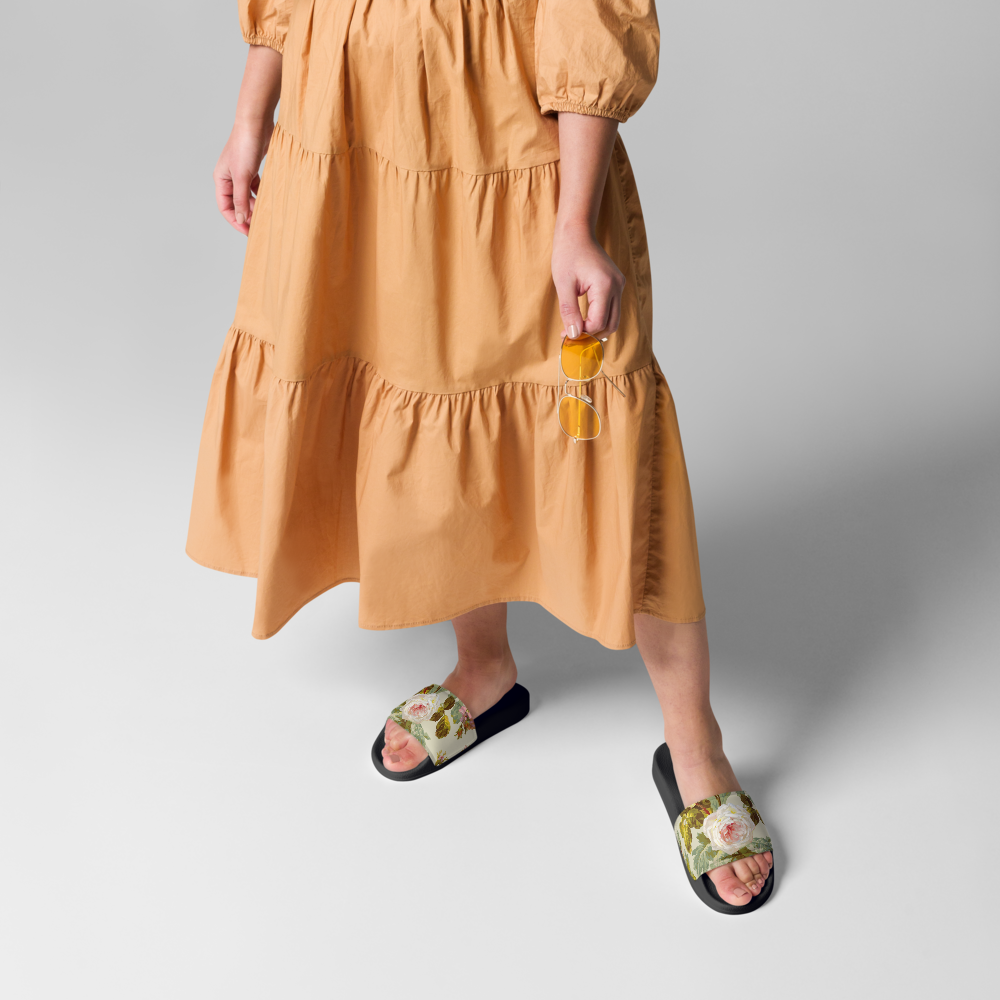 A person in an orange tiered dress holds eyeglasses and wears Women's Slides featuring floral straps and a cushioned footbed.